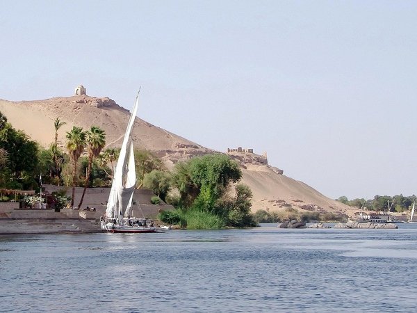 Vista del Nilo ad Aswan con una feluca, vegetazione lussureggiante e paesaggio desertico sullo sfondo.
