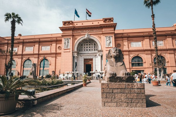The grand facade of the Egyptian Museum in Cairo, with its sphinx statue and palm-lined entrance courtyard.