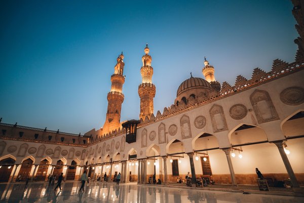 Al-Azhar Mosque's illuminated minarets and courtyard glowing at dusk against a deep blue sky.