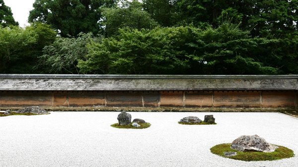 Zen rock garden of Ryoan-ji in Kyoto with rocks and white gravel, surrounded by a wall and green trees.
