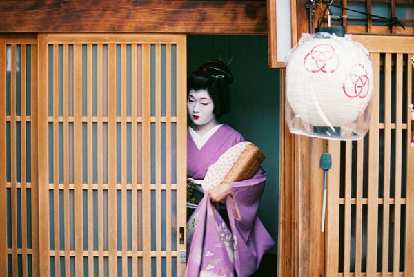 Geisha in purple kimono standing by a traditional wooden door in Kyoto's Gion District, iconic of Japanese culture.