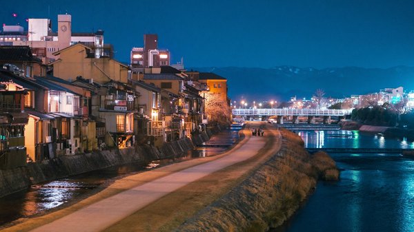 Night view of Kamo River in Kyoto with illuminated traditional houses and a bridge in the distance.