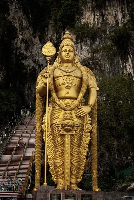 Golden statue of Lord Murugan in front of Batu Caves near Kuala Lumpur, with stairs and cliffs in the background.