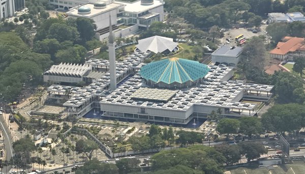 Aerial view of the National Mosque in Kuala Lumpur with its blue roof and minaret surrounded by trees and roads.