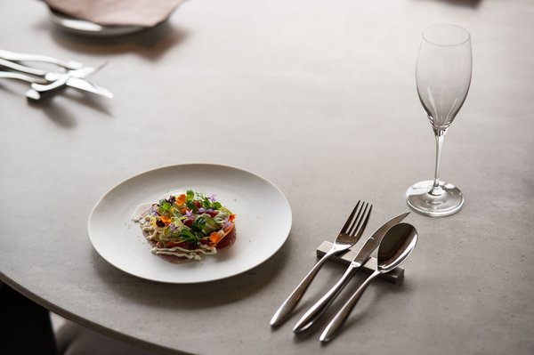 Un piatto minimalista di alta cucina guarnito con fiori edibili, servito accanto a posate lucide e un flûte da champagne.