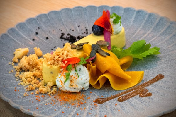 Un piatto d'alta cucina avanguardista con nastri di mango, crema alle erbe, crumble e fiori edibili disposti artisticamente su un piatto smerlato.