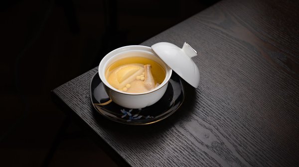 Una coscia di pollo intera cotta in un brodo dorato limpido con ginseng e giuggiole, servita in un'elegante ciotola con coperchio.