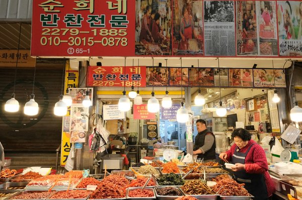 Una bancarella tradizionale di banchan coreani con vassoi metallici pieni di contorni conditi, da usare per riempire il lunchbox del mercato.
