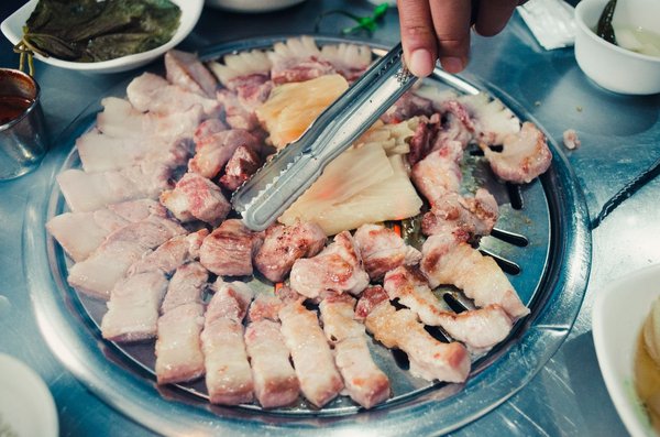 Una mano usa le pinze per girare fette di pancetta di maiale e aglio su una griglia coreana a cupola.