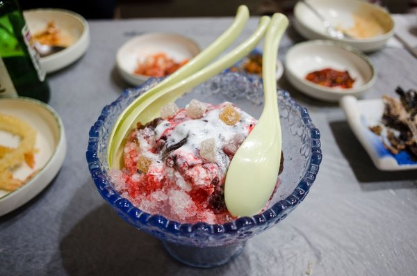 Una ciotola di patbingsu, ghiaccio grattugiato con fagioli rossi, latte condensato e tteok, servita con cucchiai lunghi.