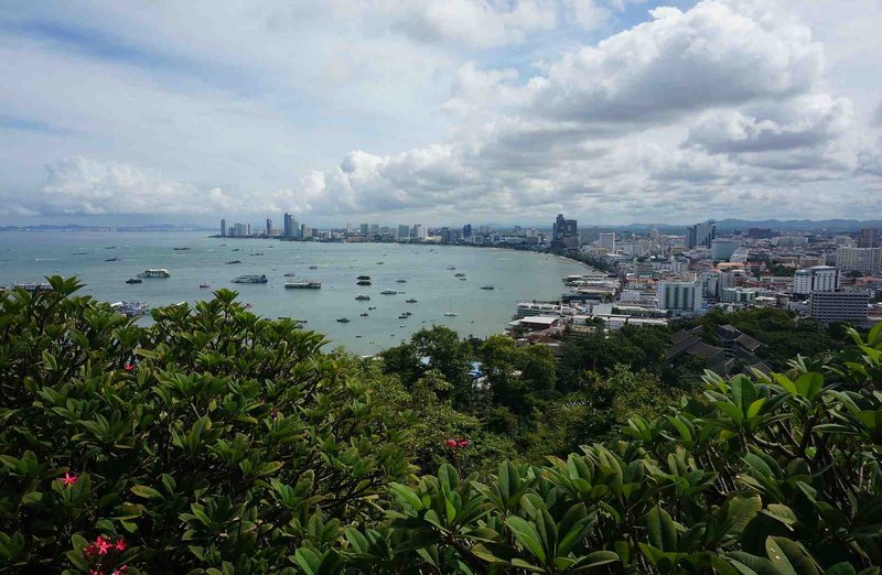 Panoramic view of Pattaya Bay with skyline, boats, and vegetation in the foreground under a partly cloudy sky.