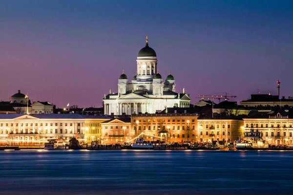 Vista notturna della Cattedrale di Helsinki illuminata, con edifici storici e riflessi sull'acqua.