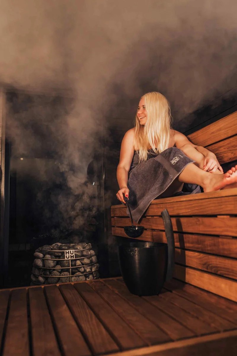 Donna seduta in una sauna tradizionale finlandese con vapore e secchio d'acqua, simbolo della cultura di Helsinki.