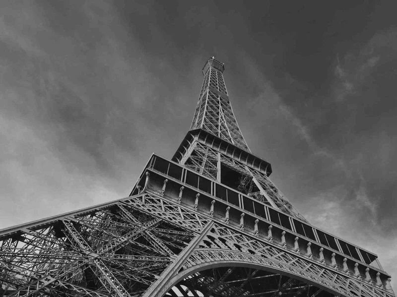 Black and white view from below the Eiffel Tower with a cloudy sky in the background.