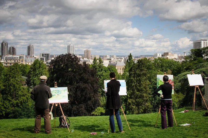 Artists painting in a park overlooking the city of Paris, cloudy sky, and green trees.