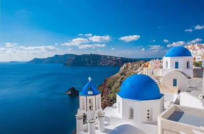 Vista panoramica di Santorini con cupole blu, mare azzurro e scogliere al tramonto.