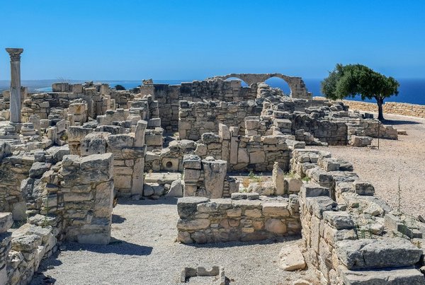 Excavated stone ruins with ancient columns and a standing archway at a Bronze Age archaeological site.