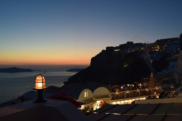 A caldera-edge restaurant terrace at sunset, tables lit with lanterns and diners overlooking the sea.