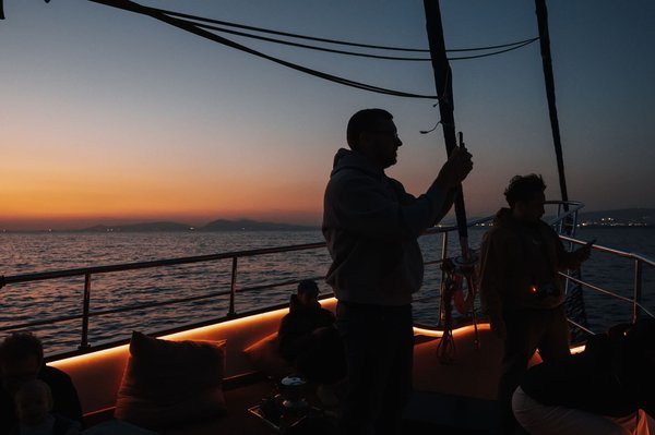 Silhouetted passengers on a catamaran's deck at sunset, warm ambient lighting glowing along the hull.