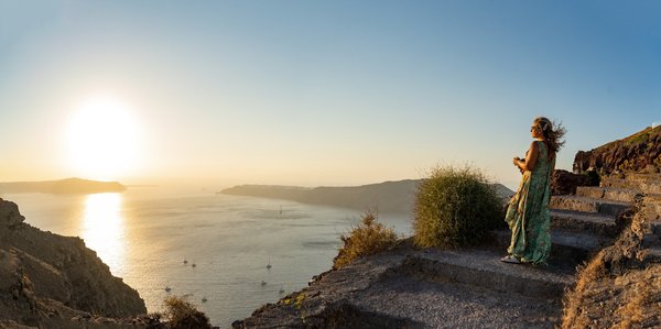 A woman pauses on the stone steps of the Fira caldera path at sunset, overlooking boats anchored below.