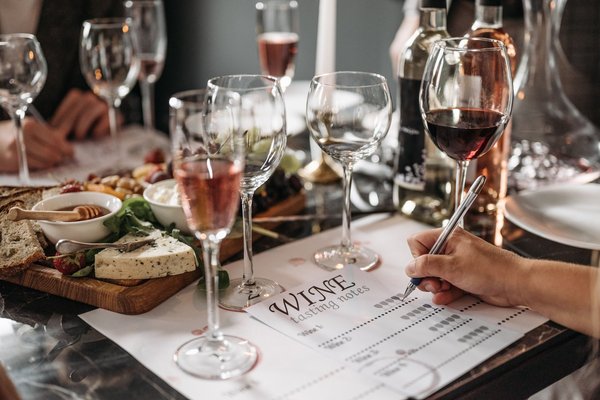A hand filling out wine tasting notes beside several glasses of red, white, and rosé wine with a cheese board.