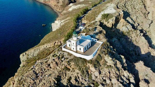 Aerial view of the Akrotiri Lighthouse in Santorini, located on a cliff with the blue sea in the background.