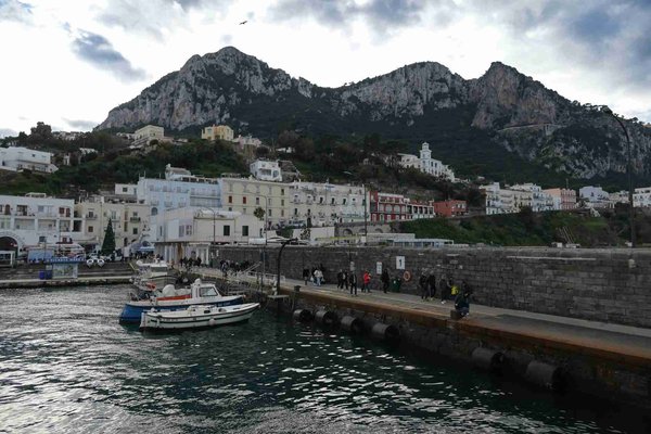 View of Capri harbor with boats, colorful buildings, and mountains in the background.