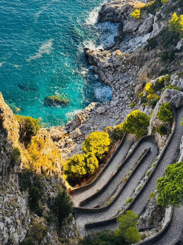 Aerial view of Via Krupp in Capri with turquoise sea and sunlit rocky cliffs.