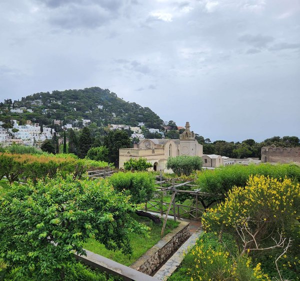 Un giardino terrazzato con arbusti in fiore e una cappella storica, con vista sulle ville bianche di Capri che scendono lungo la collina.
