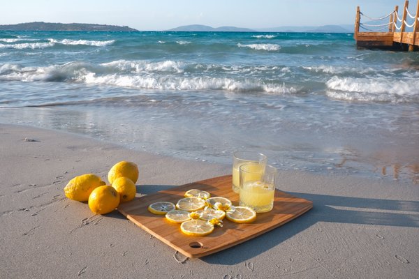 Due bicchieri di liquore al limone chiaro accanto a limoni interi e affettati su un tagliere di legno, posati sulla sabbia in riva al mare.