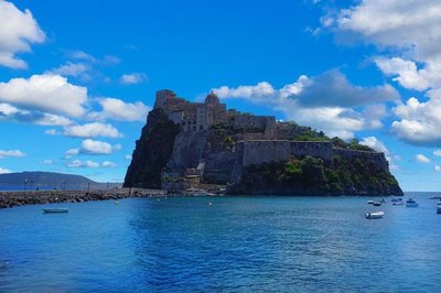 Panoramic view of the Aragonese Castle in Ischia with blue sky and boats on the sea.