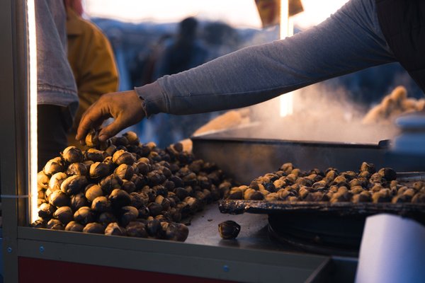 A street vendor reaches over a pile of roasted chestnuts at an outdoor cart in the evening.