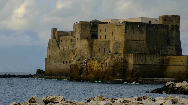 Castel dell'Ovo a Napoli con cielo nuvoloso e mare in primo piano.