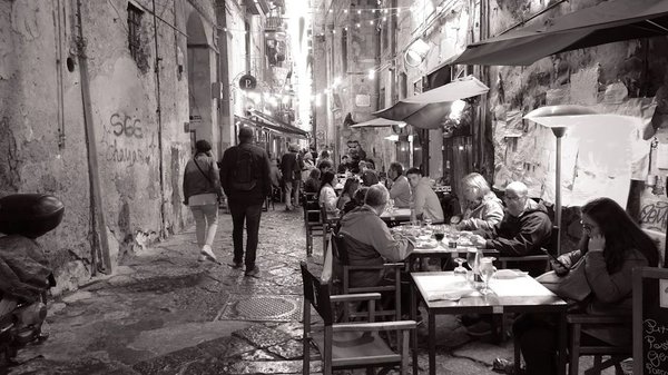 Narrow street in Naples with outdoor dining tables and people enjoying meals, lively and welcoming atmosphere.