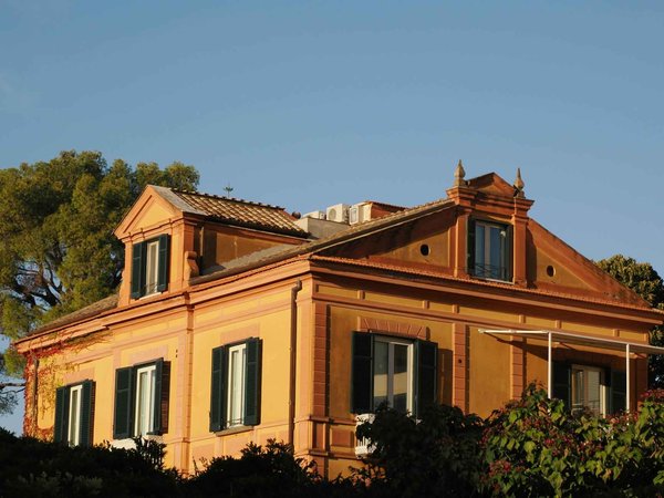 Ochre-colored villa with green shutters surrounded by greenery, lit by sunset light.