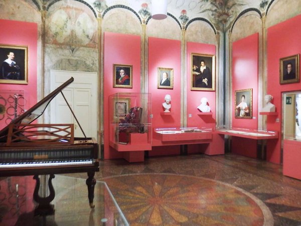 Interno del Museo della Musica di Bologna con strumenti musicali, busti e dipinti su pareti rosa.