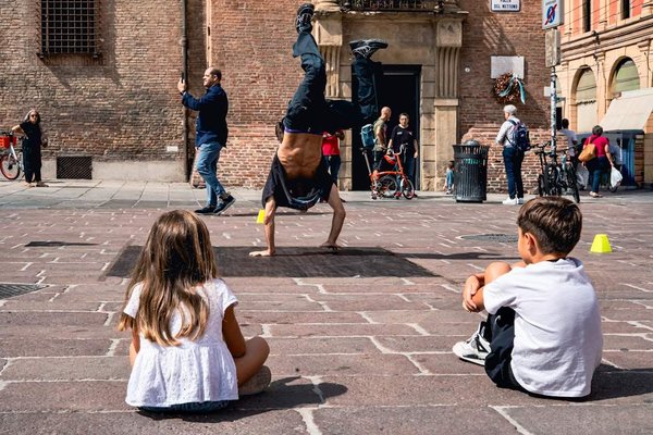 Bambini seduti su una piazza di Bologna guardano un artista di strada fare acrobazie davanti a edifici storici.