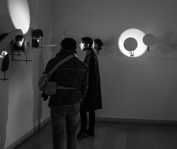 Visitors standing in a dark gallery room lit by circular mirrors and glowing spotlights.