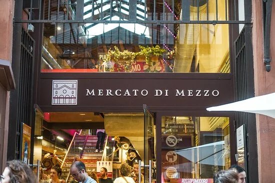 Entrance to the Mercato di Mezzo in Bologna with signage and glass windows, people entering, and visible interior decorations.