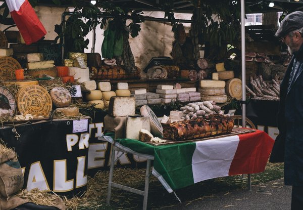Outdoor market stall with wheels of cheese and cured meats, an Italian flag draped over the table.