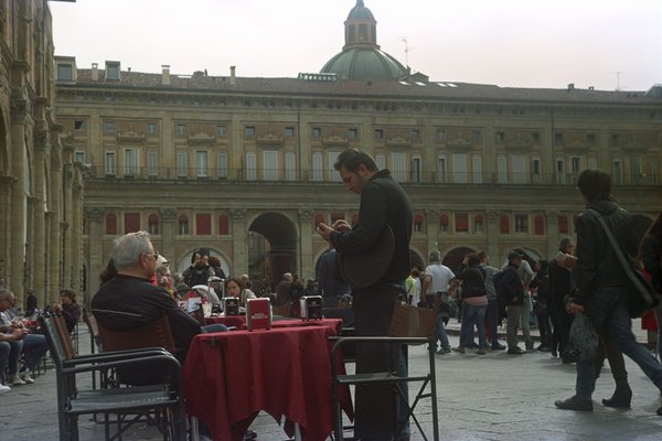 Tavoli all'aperto in una piazza con persone e un edificio storico sullo sfondo.