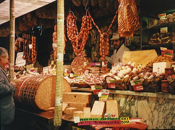 Il bancone di una tradizionale salumeria italiana con salami sospesi, un'enorme forma di mortadella e spicchi di formaggio in vendita.