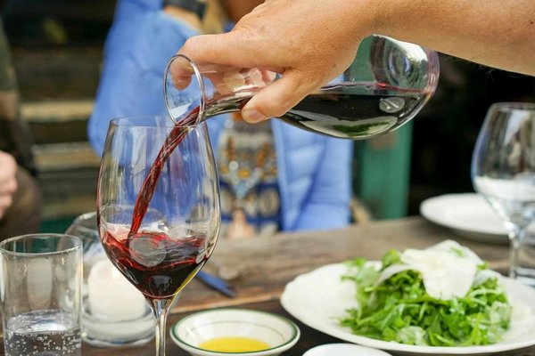 Versamento di vino rosso in un bicchiere con insalata e piatti sullo sfondo, scena di degustazione a Bologna.