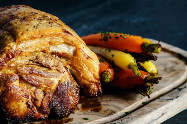 Roasted pork with crispy golden skin served with roasted baby carrots on a wooden board.