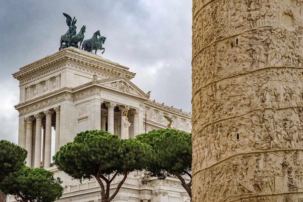 Colonna Traiana e Altare della Patria a Roma con cielo nuvoloso e pini in primo piano.