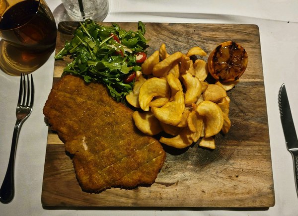 Large breaded veal cutlet on a wooden board with arugula salad and fried potato wedges.