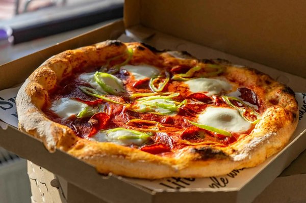 Neapolitan pizza with tomato, mozzarella, and peppers in a takeaway box, illuminated by sunlight.