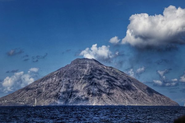 View of Stromboli volcano with blue sky and clouds, sailboats in the foreground.