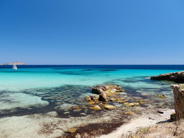 Beach with crystal-clear waters and rocks, white boat in the distance, clear blue sky.