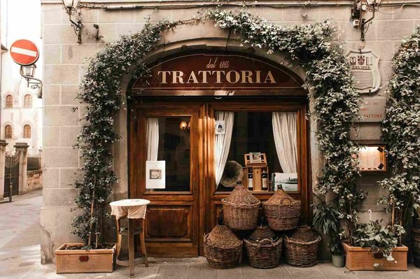 Entrance of a trattoria with 'dal 1865' sign, adorned with climbing plants and wicker baskets, in Florence.
